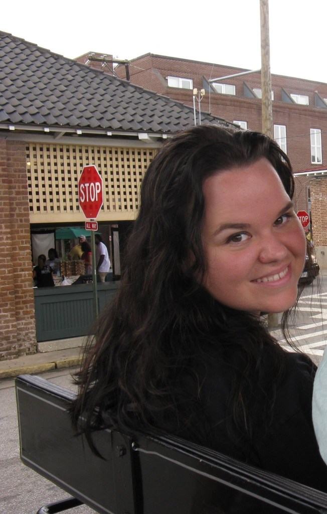 Photo of the author, white woman with long dark wavy hair looking over her right shoulder and smiling at the photographer.